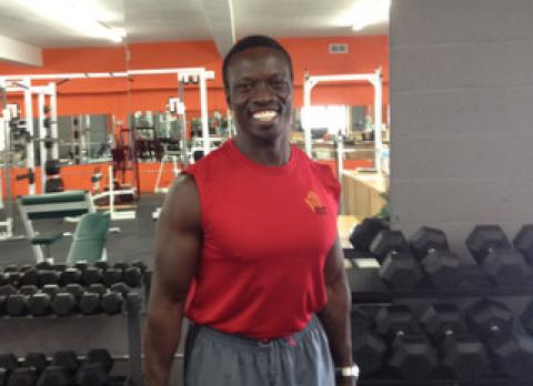 Stephen Menya standing with barbells among free weights. Stephen Menya standing with barbells among free weights.